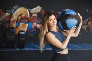 Free photo cheerful strong female sports instructor showing how workout crossfit holding medicine ball on shoulder smiling camera wear activewear workout prepare squats training equipment